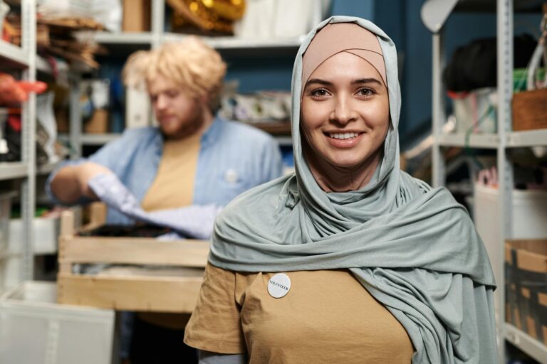 Smiling woman in hijab volunteering in a storeroom with a colleague organizing supplies.